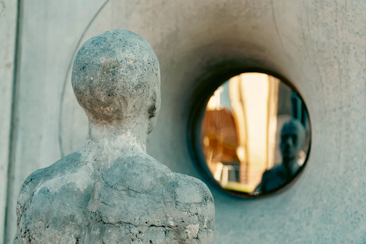 A concrete figure looking at its own reflection in a round mirror set into a wall.