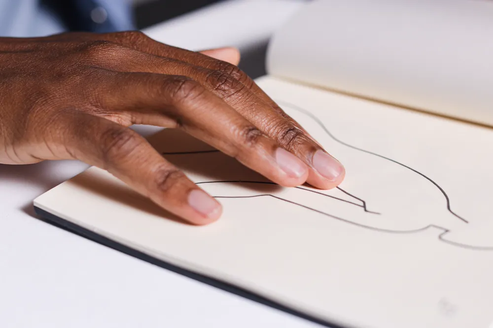 A hand resting on an open sketchbook with a single line drawing in progress.